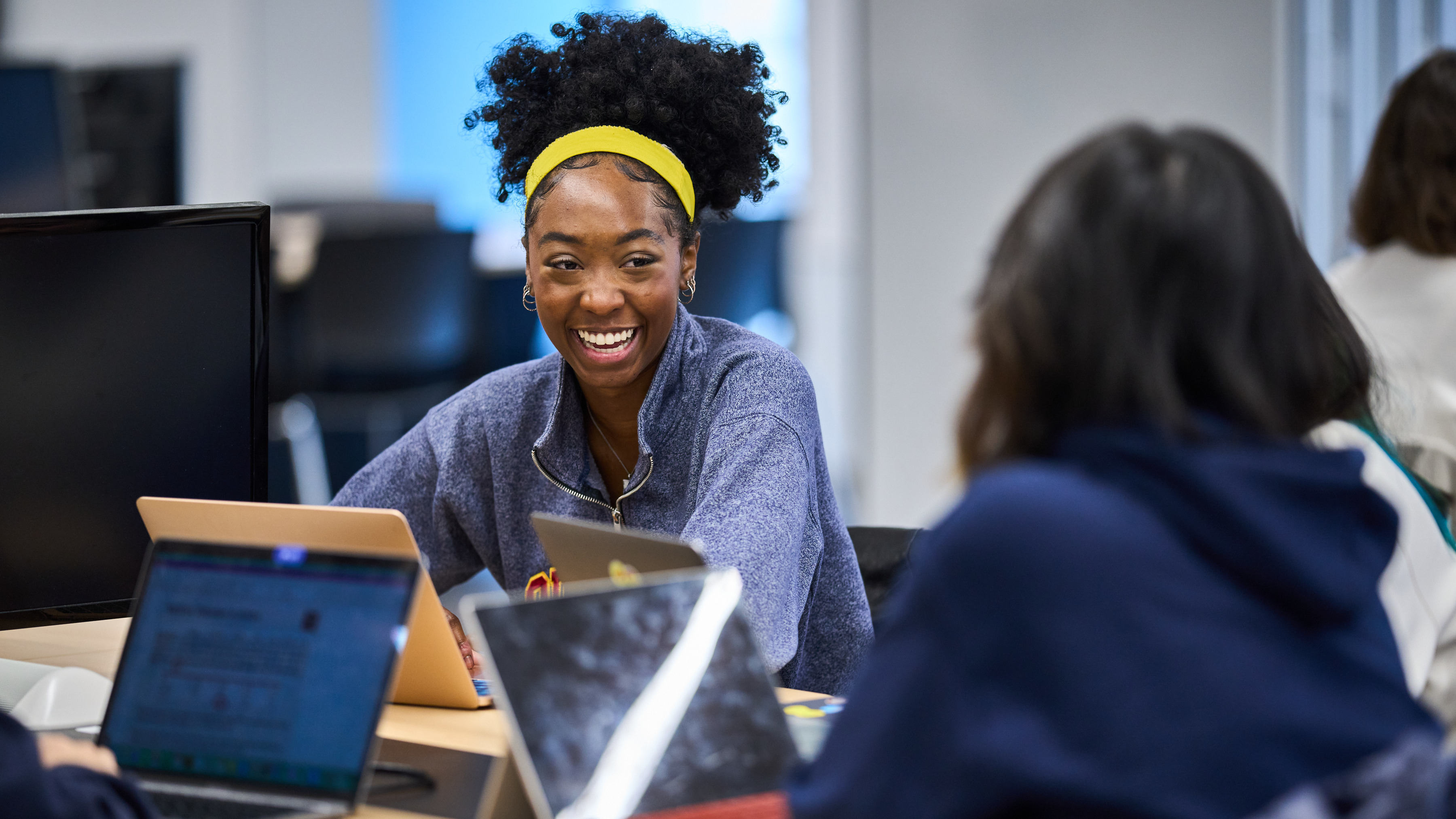 Students work together on laptops in a classroom while one student smiles during the discussion.